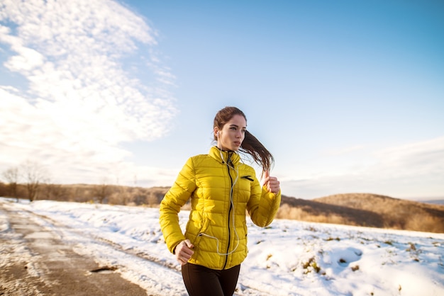 junges motiviertes und fokussiertes sportliches aktives Mädchen mit einem flauschigen Pferdeschwanz in der Wintersportbekleidung, die in der verschneiten Natur auf der Straße läuft.