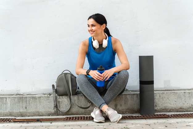 Junges, müdes, sportliches Mädchen, das eine Pause vom Training macht, während es eine blaue Flasche hält und in der Nähe der grauen Wand an der sonnigen Morgenstadtstraße im Freien sitzt. Stock Foto