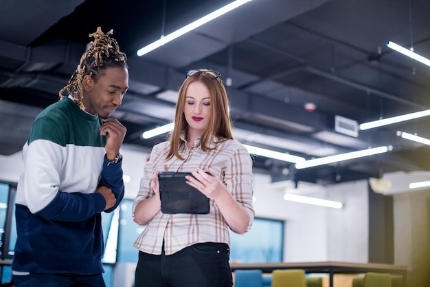 Junges multiethnisches Geschäftspaar Afroamerikaner und seine rothaarige Kollegin benutzen einen Tablet-Computer, während sie in einem modernen Startup-Büro stehen