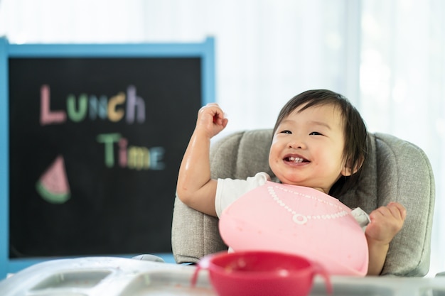 Junges nettes Kind auf dem Fütterungssitz des Babyhochstuhls, der zu Hause Wassermelone mit Lächelngesicht hält, genießen, süße Frucht der Mahlzeit zu essen und Glück zu lachen.