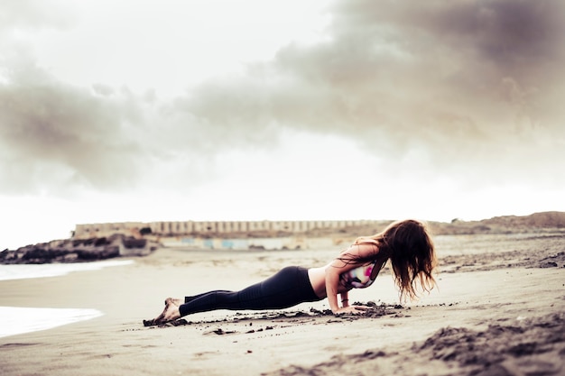 Junges, nicht erkennbares, attraktives brünettes Mädchen, das Plank Pilates Position am Strand am Ufer dopingt - Sport in der Natur im Freien genießen - warmer Filter und bewölkter Himmel