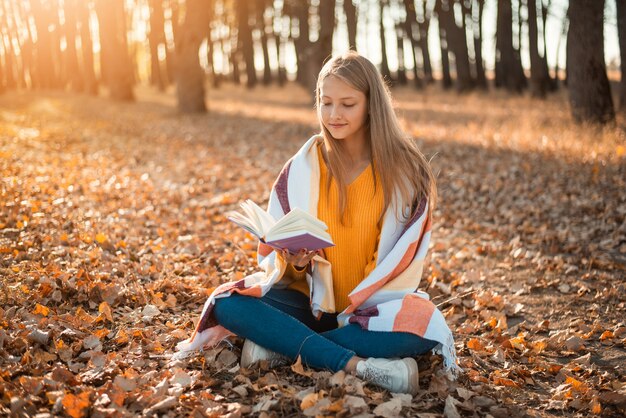 Junges niedliches Schulmädchen, das im Plaid im Herbstwald sitzt und ein Buch liest