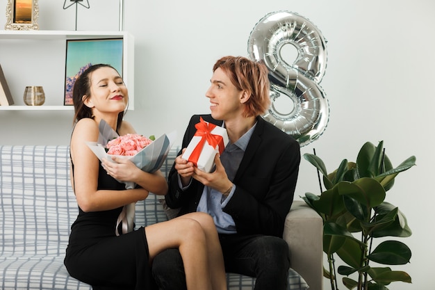 Junges Paar am glücklichen Frauentag, der Geschenk mit Blumenstrauß auf dem Sofa im Wohnzimmer hält