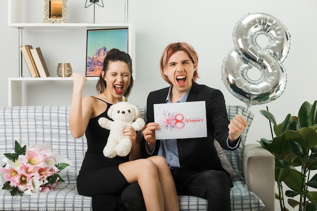 Junges Paar am glücklichen Frauentag mit Teddybär und Postkarte auf dem Sofa im Wohnzimmer sitzend