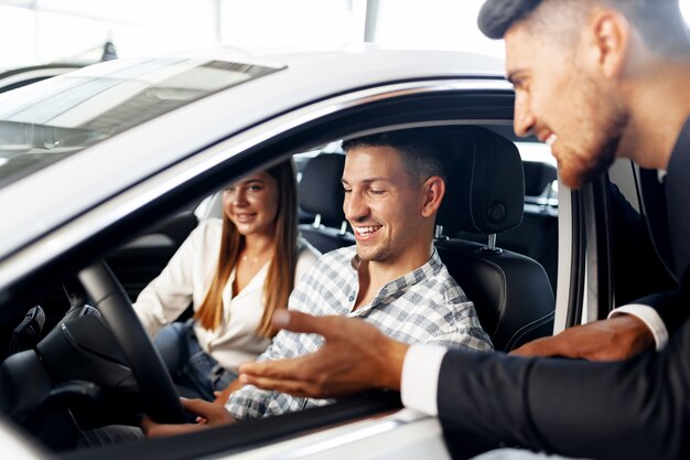 Junges Paar, das ein Auto im Autohaus wählt, mit Manager, der ihnen hilft