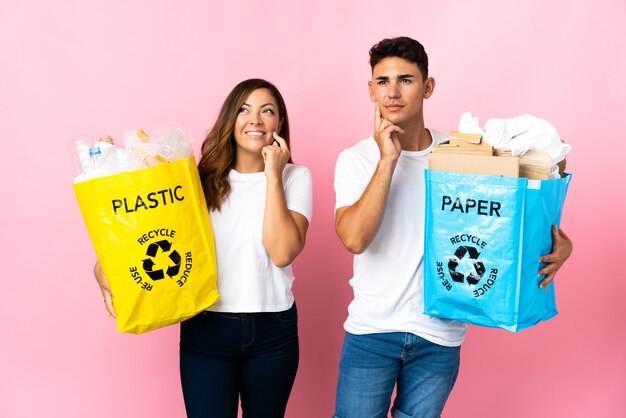 Junges Paar, das eine Tasche voll Plastik und Papier auf Rosa hält und eine Idee beim Aufschauen denkt