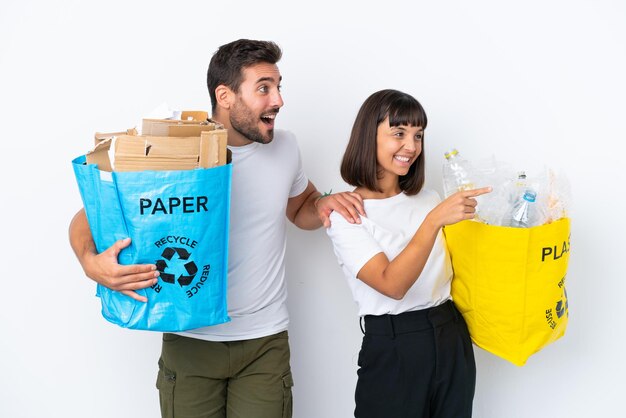 Foto junges paar, das eine tasche voller plastik und papier zum recycling hält, isoliert auf weißem hintergrund, das auf die seite zeigt, um ein produkt zu präsentieren