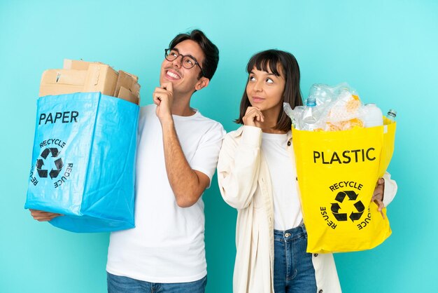 Junges Paar, das eine Tüte voller Plastik und Papier hält, um es einzeln auf blauem Hintergrund zu recyceln und eine Idee zu denken, während es nach oben schaut