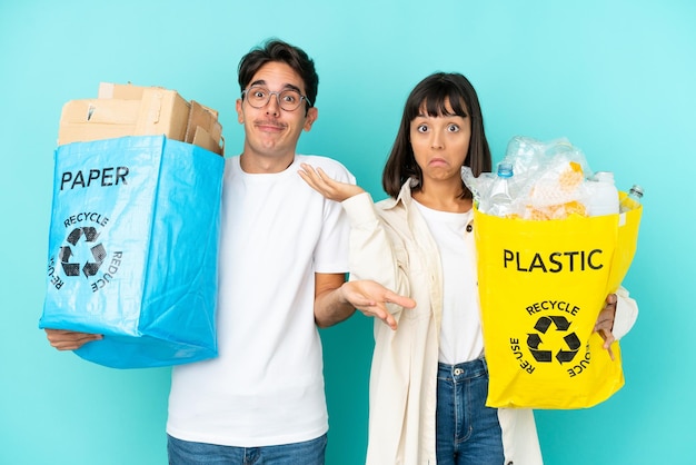 Junges Paar, das eine Tüte voller Plastik und Papier hält, um es isoliert auf blauem Hintergrund zu recyceln, und Zweifel hat, während es Hände und Schultern hebt