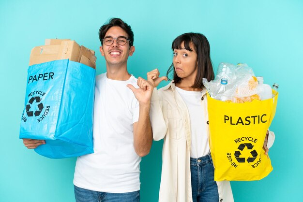 Junges Paar, das eine Tüte voller Plastik und Papier hält, um sie isoliert auf blauem Hintergrund zu recyceln, stolz und selbstzufrieden im Konzept der Liebe