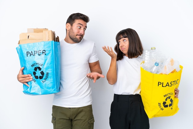 Junges Paar, das eine Tüte voller Plastik und Papier hält, um sie isoliert auf weißem Hintergrund zu recyceln, und dabei eine unwichtige Geste macht, während es die Schultern anhebt