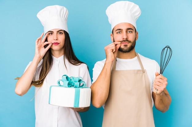 Junges Paar, das einen Kuchen zusammen isoliert mit den Fingern auf den Lippen kocht, die ein Geheimnis halten.