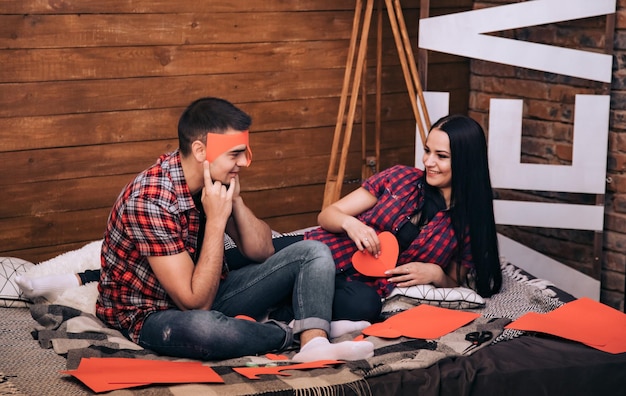 Junges Paar, das Herzen aus rotem Papier zum Valentinstag macht, sitzt auf dem Bett und lacht zusammen über den hölzernen Hintergrund Romantik- und Liebeskonzept