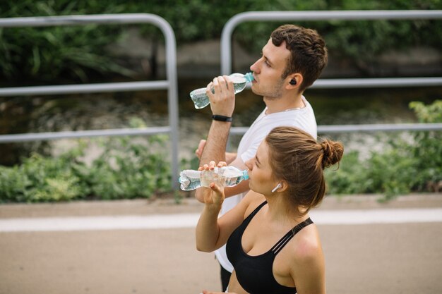 Junges Paar, das im Stadtpark mit Plastikflasche Wasser in den Händen läuft, gemeinsamer Sport, Fröhlichkeit, Stadtsport gesunder Lebensstil, Fitness zusammen, Sommerabend, Läufer, Trinkwasser, Durst