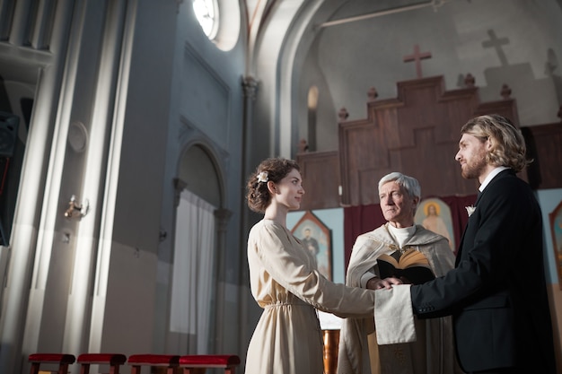 Junges Paar, das in der Kirche heiratet, während der Priester die Gebete liest