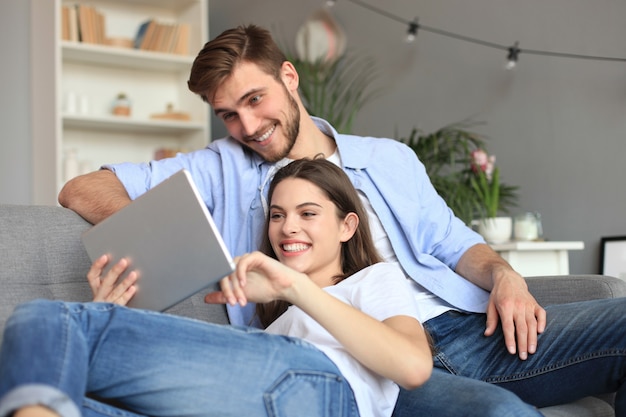 Junges Paar, das Medieninhalte online in einem Tablet ansieht, das auf einem Sofa im Wohnzimmer sitzt.