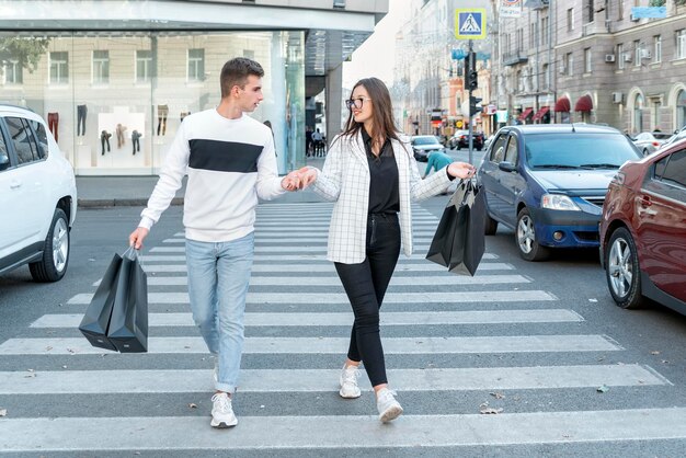 Junges Paar, das nach dem Einkaufen zusammen über die Straße geht Mann und Frau mit schwarzen Taschen mit Einkäufen