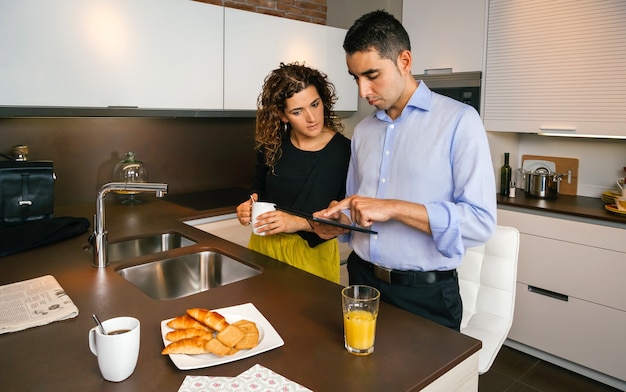 Junges Paar, das Nachrichten in elektronischer Tablette sucht, während es schnellen Kaffee zu Hause trinkt, bevor es zur Arbeit geht