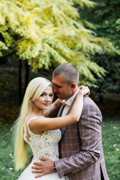 Junges Paar, das romantische Momente beim Gehen im Park genießt. Stilvolle Braut und Bräutigam, die an ihrem Hochzeitstag im Park posieren und küssen. Elegante Braut im schönen weißen Kleid, Bräutigam im Anzug.