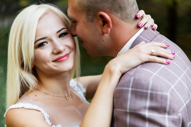 Junges Paar, das romantische Momente beim Gehen im Park genießt. Stilvolle Braut und Bräutigam, die an ihrem Hochzeitstag im Park posieren und küssen. Elegante Braut im schönen weißen Kleid, Bräutigam im Anzug.