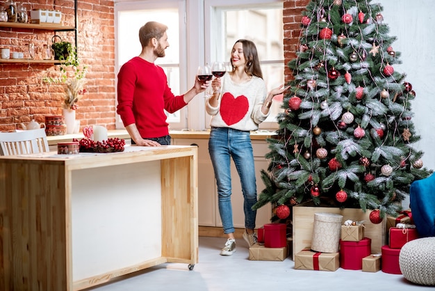 Junges Paar, das Spaß hat, mit Weingläsern zu stehen und Neujahrsferien in der Nähe des Weihnachtsbaums in ihrem Haus zu feiern?