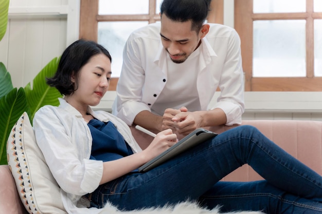 Junges Paar, das zu Hause zusammenarbeitet. Frau sitzt auf dem Sofa mit Tablet und Mann beobachtet sie bei der Arbeit. Kreative Bürokollegen diskutieren die neue Idee und das Storyboard im Wohnzimmer.