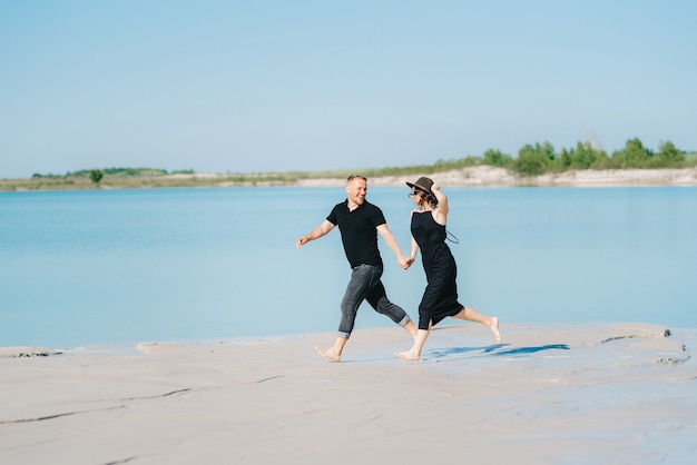 Junges Paar, ein Mann mit einem Mädchen in schwarzer Kleidung, geht auf dem weißen Sand am Rand des blauen Wassers spazieren