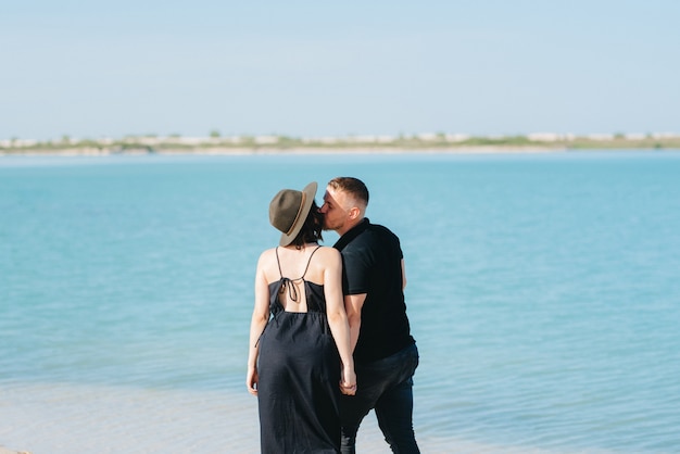 Junges Paar, ein Mann mit einem Mädchen in schwarzer Kleidung, geht auf dem weißen Sand am Rand des blauen Wassers spazieren