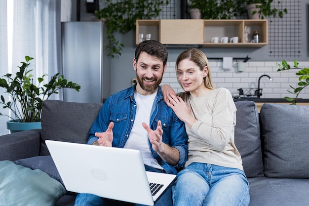 Junges Paar, Familienvater und -frau, die zu Hause auf der Couch sitzen und über Videoanruf vom Laptop mit Freunden der Familie sprechen. Sie winken, lächeln, grüßen