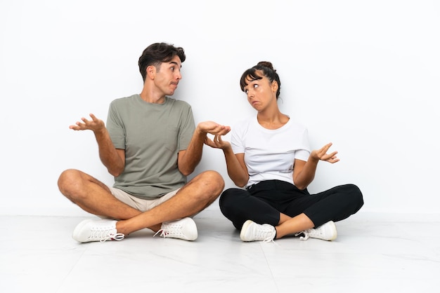 Junges Paar gemischter Rassen, das isoliert auf weißem Hintergrund auf dem Boden sitzt und beim Anheben der Schultern eine unwichtige Geste macht