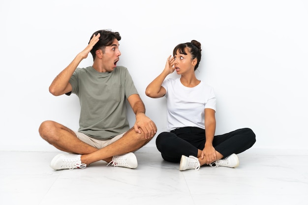 Junges Paar gemischter Rassen, das isoliert auf weißem Hintergrund mit überraschtem und schockiertem Gesichtsausdruck auf dem Boden sitzt