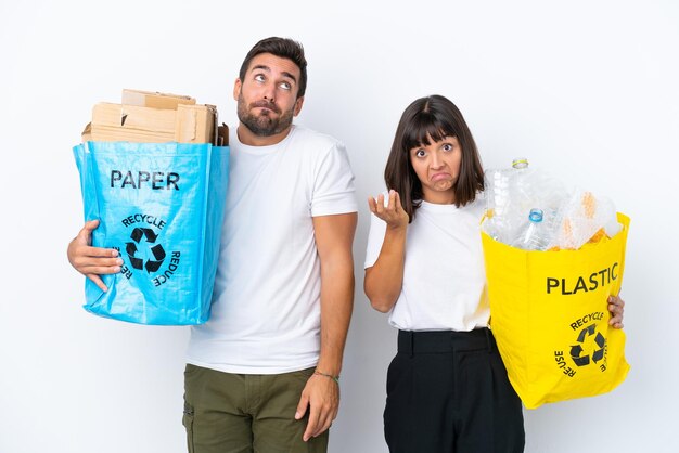 Junges Paar hält eine Tüte voller Plastik und Papier, um sie isoliert auf weißem Hintergrund zu recyceln, und macht Zweifel, während es die Schultern anhebt