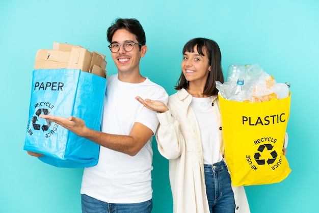 Junges Paar hält eine Tüte voller Plastik und Papier zum Recyceln isoliert auf blauem Hintergrund und streckt die Hände zur Seite, um zum Kommen einzuladen