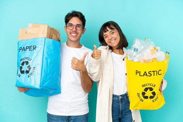 Junges Paar hält eine Tüte voller Plastik und Papier zum Recyceln isoliert auf blauem Hintergrund und zeigt mit beiden Händen eine Daumen-hoch-Geste und lächelt