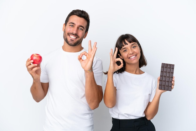 Junges Paar hält Schokolade und Apfel isoliert auf weißem Hintergrund und zeigt ein Ok-Zeichen mit den Fingern