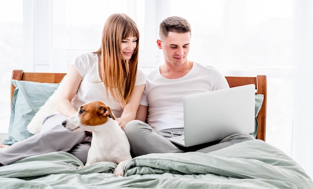 Junges Paar im Bett mit Geräten und süßem Hund. Schöne Frau und Mann, die Laptop und Mädchen betrachten, die Smartphone halten. Familienmorgen mit Haustier und Technologien