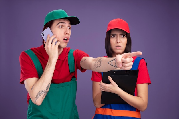 Junges Paar in Bauarbeiteruniform und Mütze mit Blick auf die Seite überraschter Kerl, der am Telefon spricht und auf die Seite zeigt verwirrtes Mädchen mit Zwischenablage