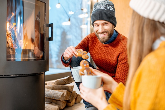 Junges Paar in hellen Pullovern mit heißen Getränken am Kamin im modernen Haus in den Bergen während der Winterzeit?