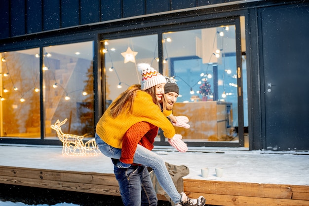 Junges Paar in hellen Pullovern und Hüten, die sich während der Winterferien in der Nähe des dekorierten Hauses amüsieren