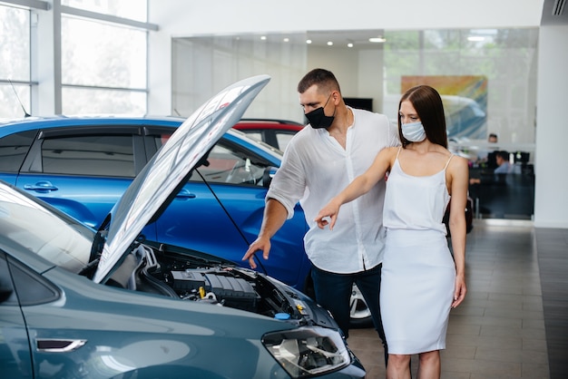 Junges Paar in Masken wählt ein neues Fahrzeug aus und konsultiert einen Vertreter des Autohauses in der Zeit der Pandemie.