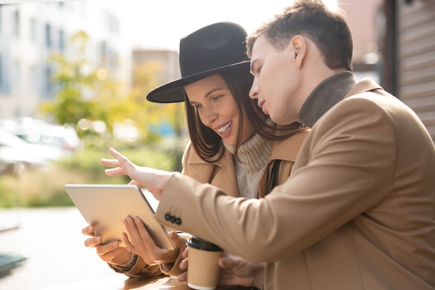 Junges Paar in stilvoller Freizeitkleidung, das sich im Café im Freien ausruht und über Online-Informationen diskutiert, während der Mann auf das Tablet-Display zeigt?