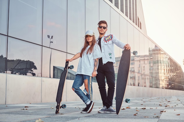 Junges Paar in trendiger Kleidung posiert mit Skateboards in der Nähe von Wolkenkratzern.