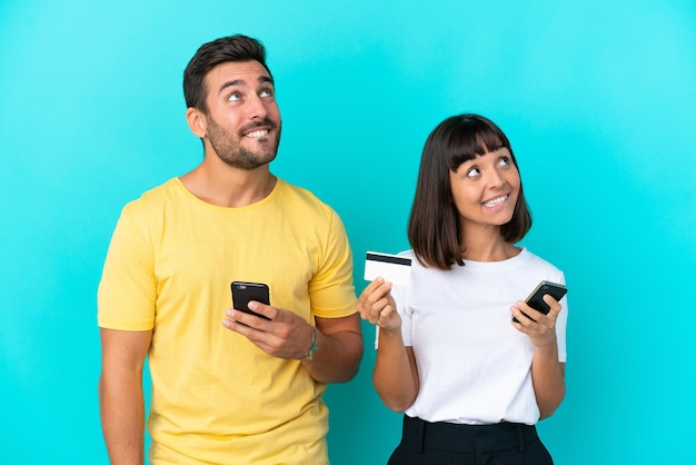 Junges Paar isoliert auf blauem Hintergrund, das beim Denken mit dem Handy mit einer Kreditkarte kauft