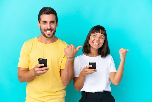 Junges Paar isoliert auf blauem Hintergrund mit Handy und zeigt auf die Seite