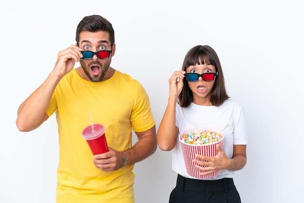 Junges Paar isoliert auf weißem Hintergrund überrascht mit 3D-Brille und hält einen großen Eimer Popcorn