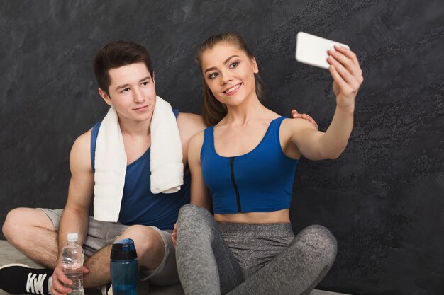Junges paar macht selfie im fitnessstudio, sitzt an der wand. sportler, mann und frau, die nach dem training eine pause auf dem smartphone machen, kopieren raum
