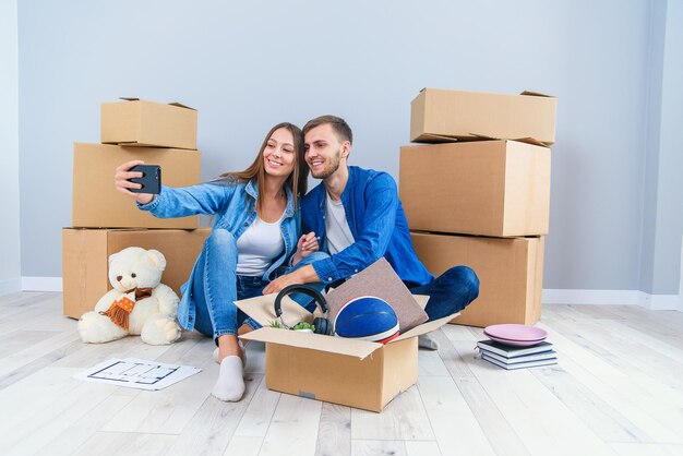 Junges Paar macht Selfies in ihrem neuen Zuhause, während es unter Kartonkisten sitzt, glücklich und attraktiv