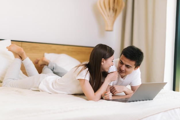 Junges Paar Mädchen und Kerl liegen auf dem Bett vor dem Laptop im hellen Schlafzimmer. Hübsche junge Frau flirtet mit ihrem Freund und kitzelt sein Gesicht sanft mit ihren Haaren im hellen Schlafzimmer