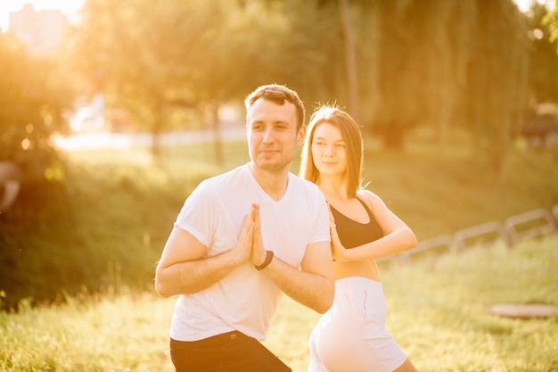 Junges Paar, Mann und Frau, die Sport treiben, Yoga auf dem Rasen der Stadt, Sommerabend, sich bei Sonnenuntergang zusammenstrecken, Konzentration auf den Sonnenuntergang