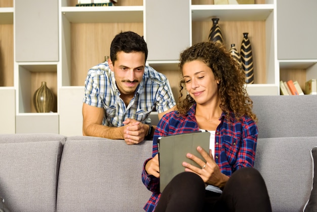 Junges Paar mit einem Tablet-PC, der zusammen sitzt und es mit erfreutem Lächeln in ihrem Wohnzimmer beobachtet